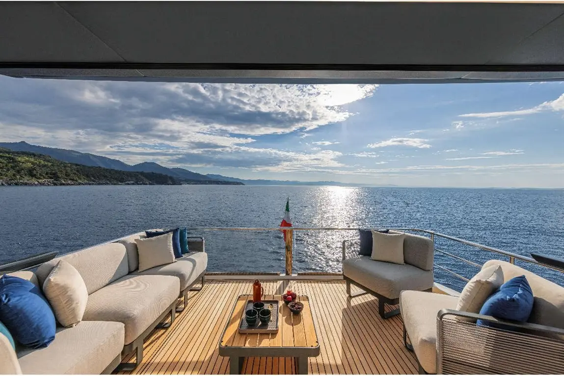 Luxurious yacht terrace with seating, a table set with drinks, overlooking the tranquil sea and distant mountains under a blue sky.